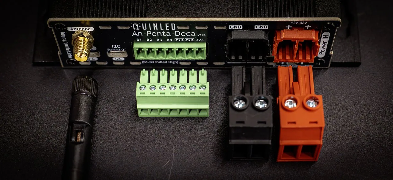 QuinLED An-Penta-Deca top panel close-up with detached green 8-pin and orange/black power terminals and antenna laid in front on a dark mat