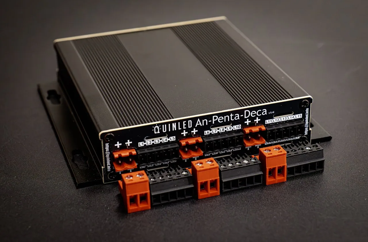 QuinLED An-Penta-Deca 15-channel dimmer at an angle, finned aluminium case with orange and black L1-L15 plug-in screw terminals on a dark surface