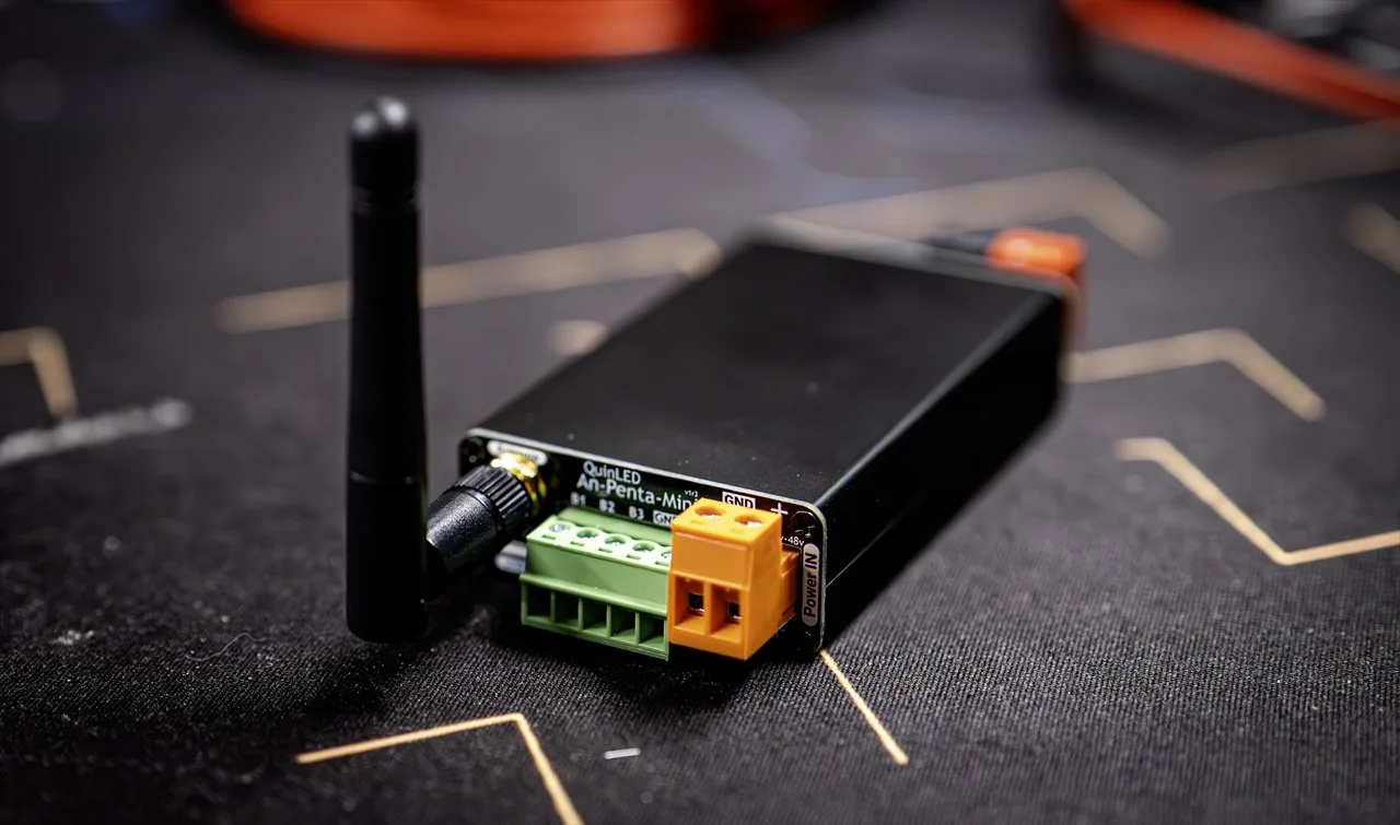 QuinLED An-Penta-Mini angled on a dark mat, green output and orange Power IN terminals fitted, WiFi antenna upright, blurred circuit backdrop