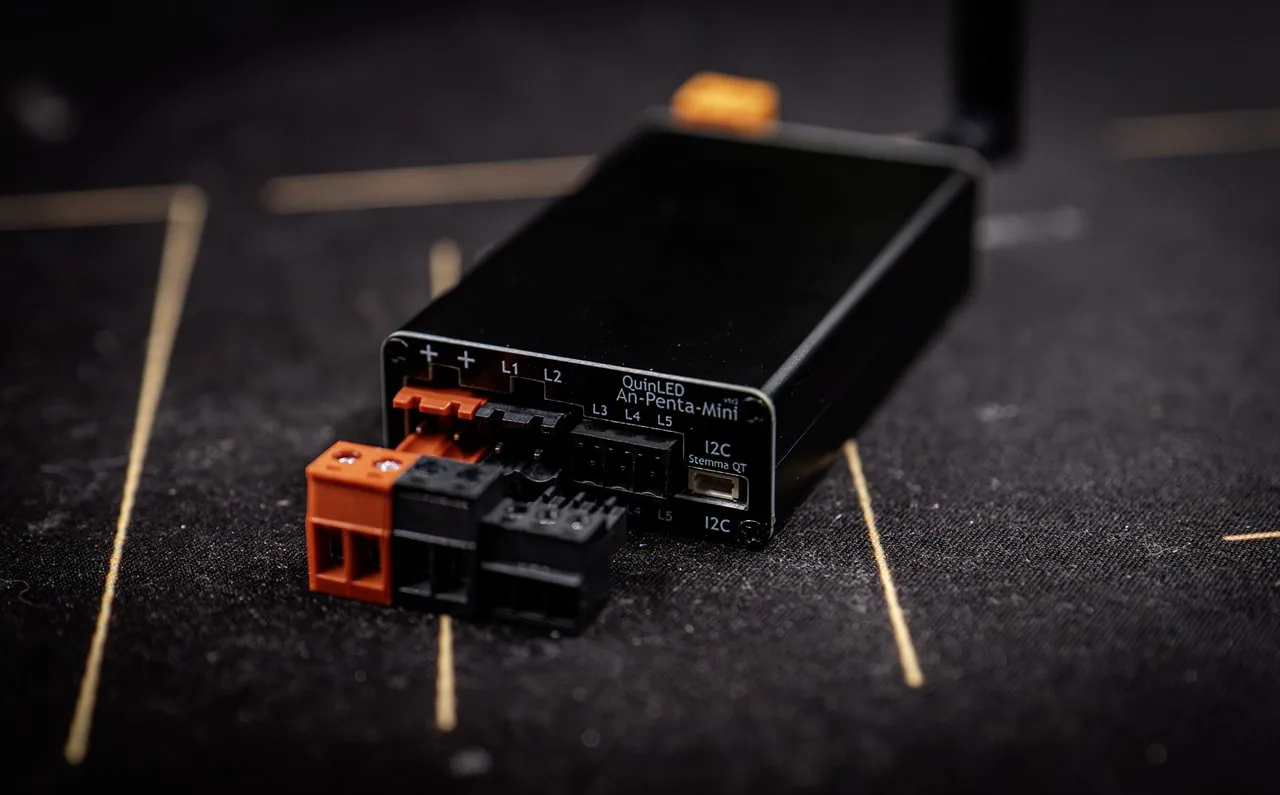 QuinLED An-Penta-Mini at an angle on a dark mat, detachable orange and black terminal blocks in front, antenna tilted back, L1-L5 labels