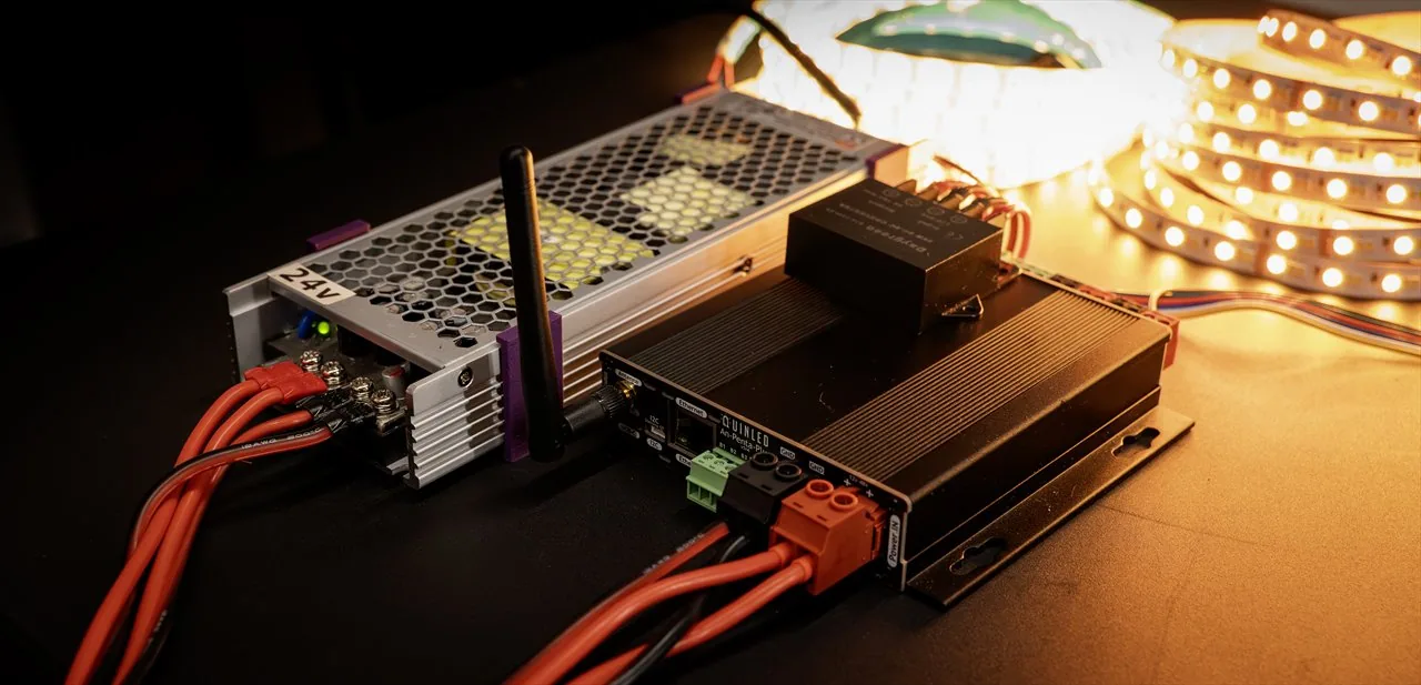 QuinLED An-Penta-Plus finned case beside a 24V power supply with antenna, wired to warm-white LED strip reels glowing in the dark