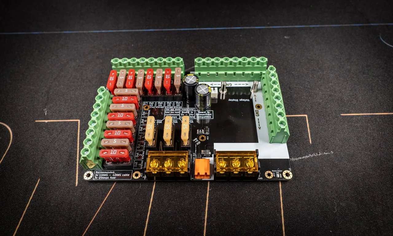 QuinLED Dig-Octa Power-7 at an angle, black Powerboard ringed with green screw terminals, blade fuses and gold bolt inputs on a dark mat